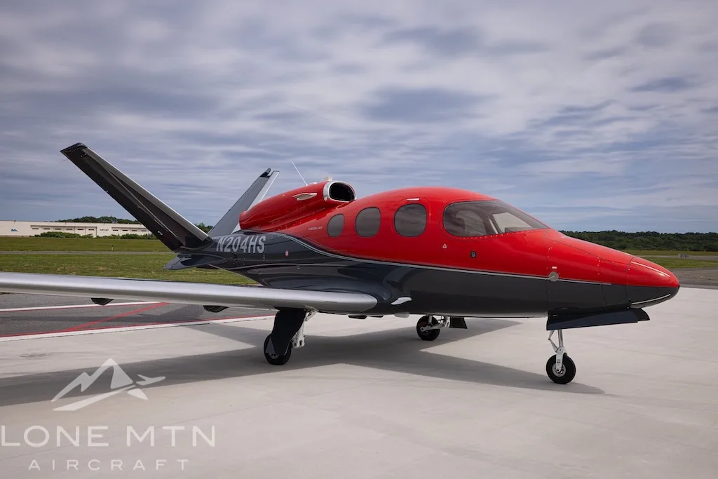 A Cirrus Vision Jet N204HS in red and grey paint scheme, parked on an airfield under a cloudy sky.