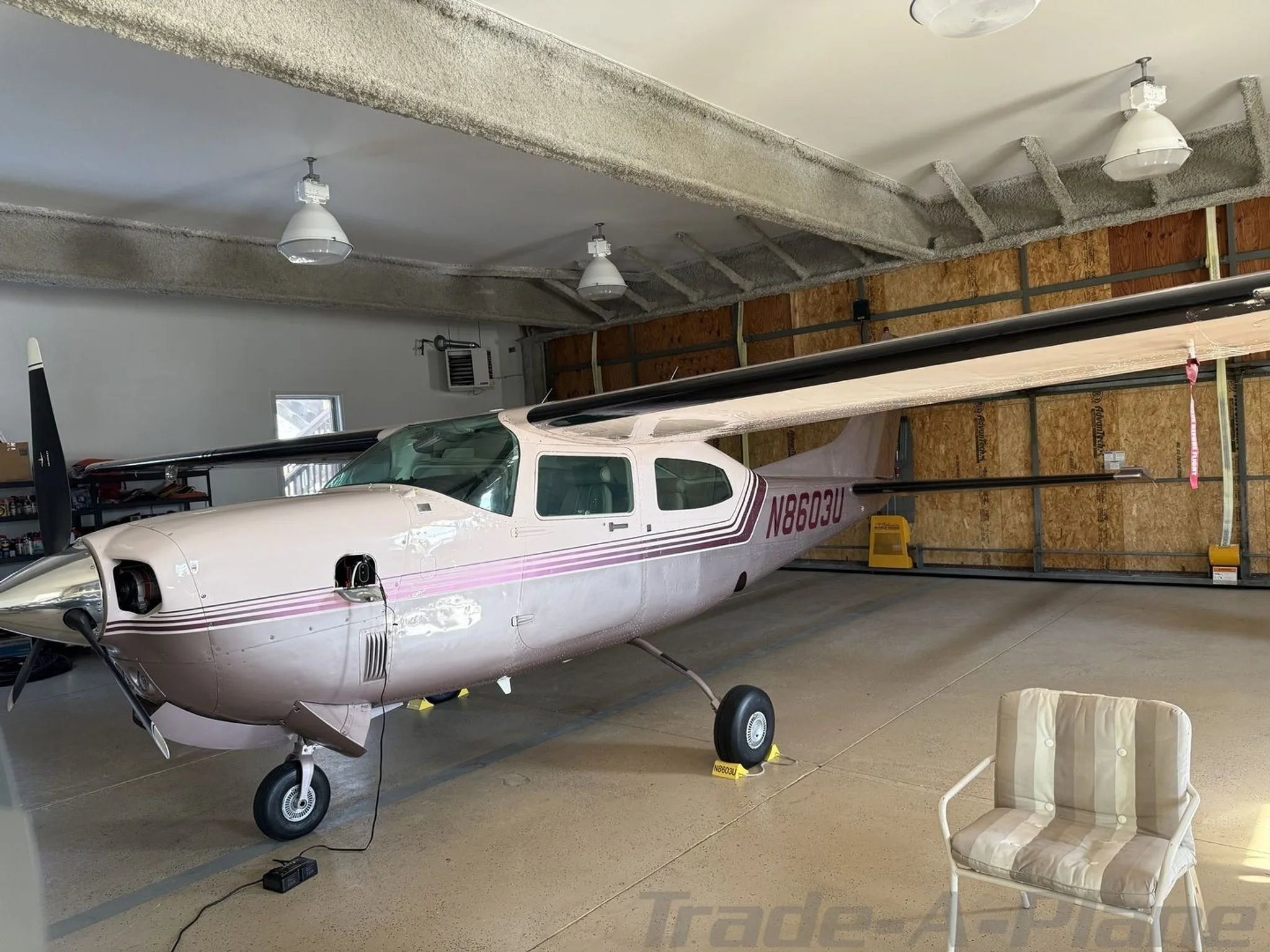A front-side view of a light pink and purple Cessna 210 Centurion aircraft, registration N8603U, parked in a hangar.