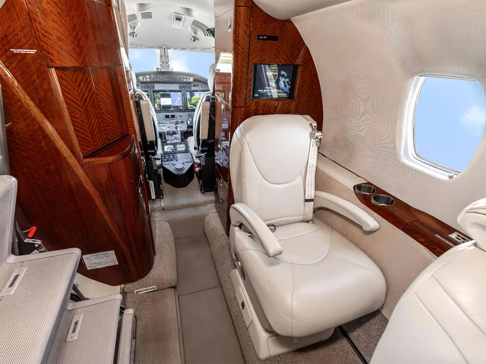 A private jet cabin interior showing a single light leather seat, wood paneling, and a view into the cockpit.