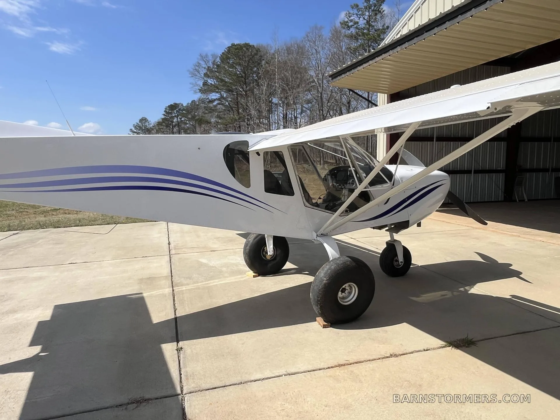 A white high-wing aircraft with blue stripes, parked on a concrete pad next to an open hangar.
