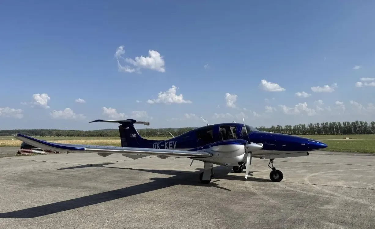 A blue and silver Diamond DA62 aircraft with registration OK-KEV is parked on a tarmac under a blue sky.