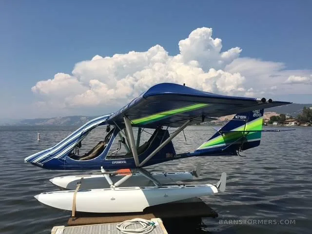 A blue and green experimental seaplane with white floats is docked on a calm lake under a partly cloudy sky.