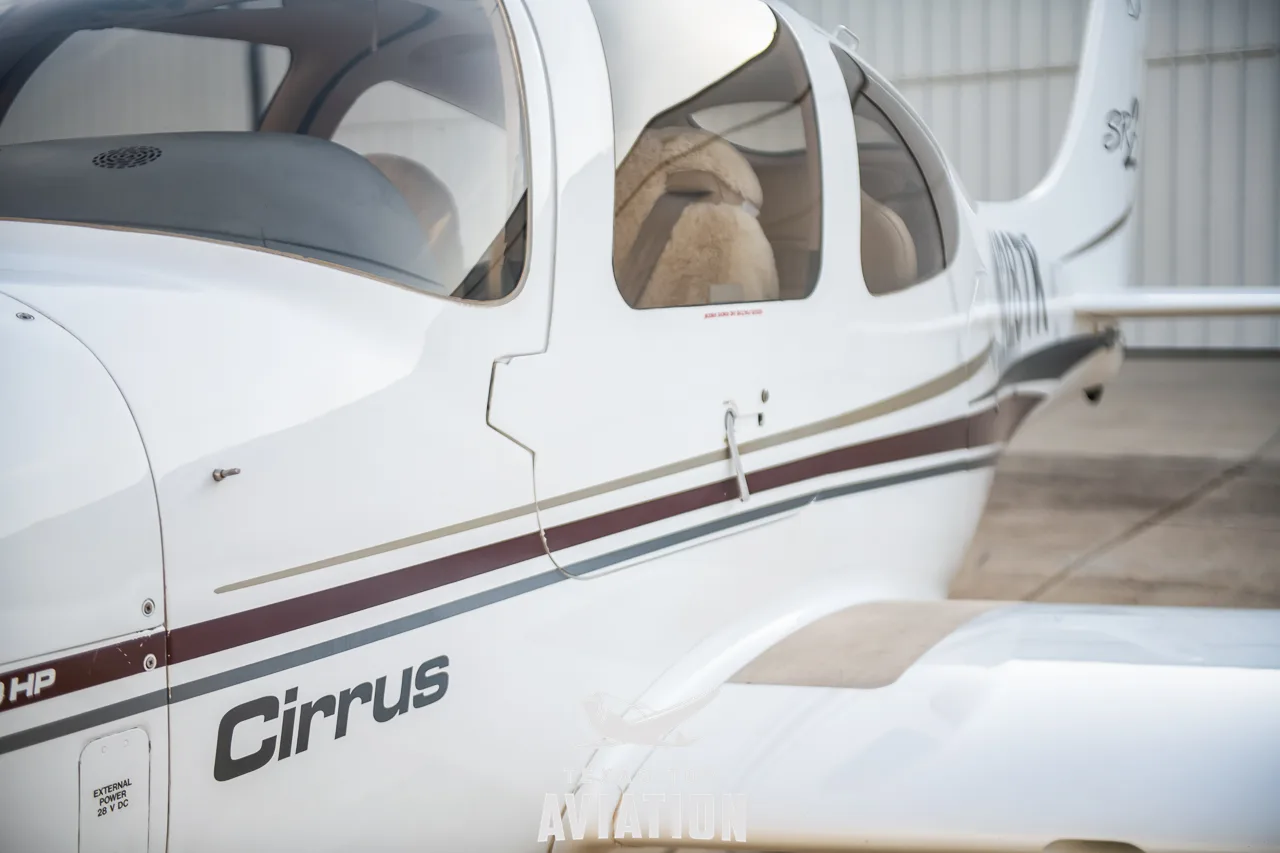 A close-up view of the white fuselage of a Cirrus SR22 aircraft, showing maroon and gold stripes and the "Cirrus" logo.