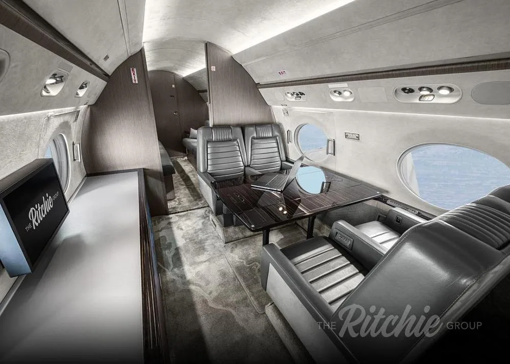 A view inside a private jet cabin showing grey leather seating, a dark wood table with a laptop, and a flat-screen monitor.