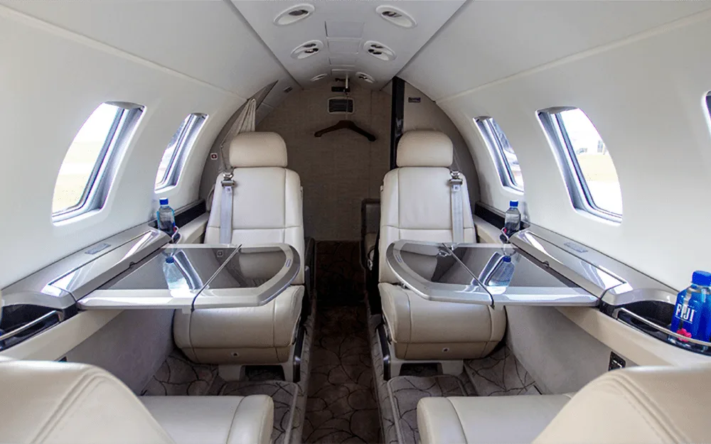 The aircraft cabin shows two rows of light-colored leather seats with fold-out tables and Fiji water bottles.