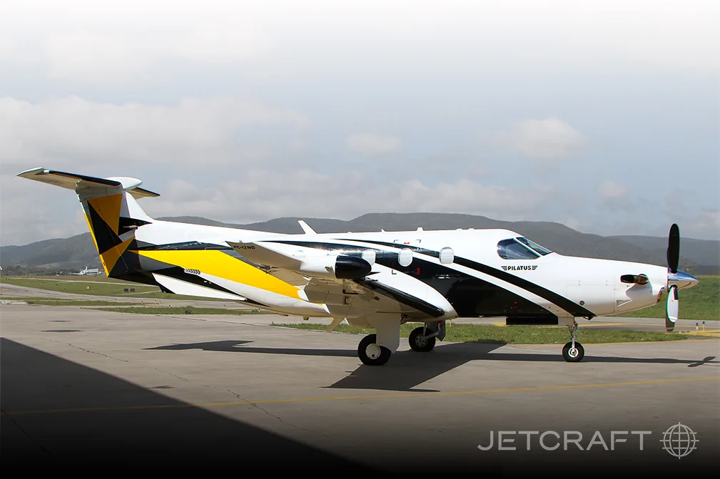 A white, black, and yellow Pilatus PC-12 NGX turboprop aircraft is parked on an airport tarmac under a partly cloudy sky.