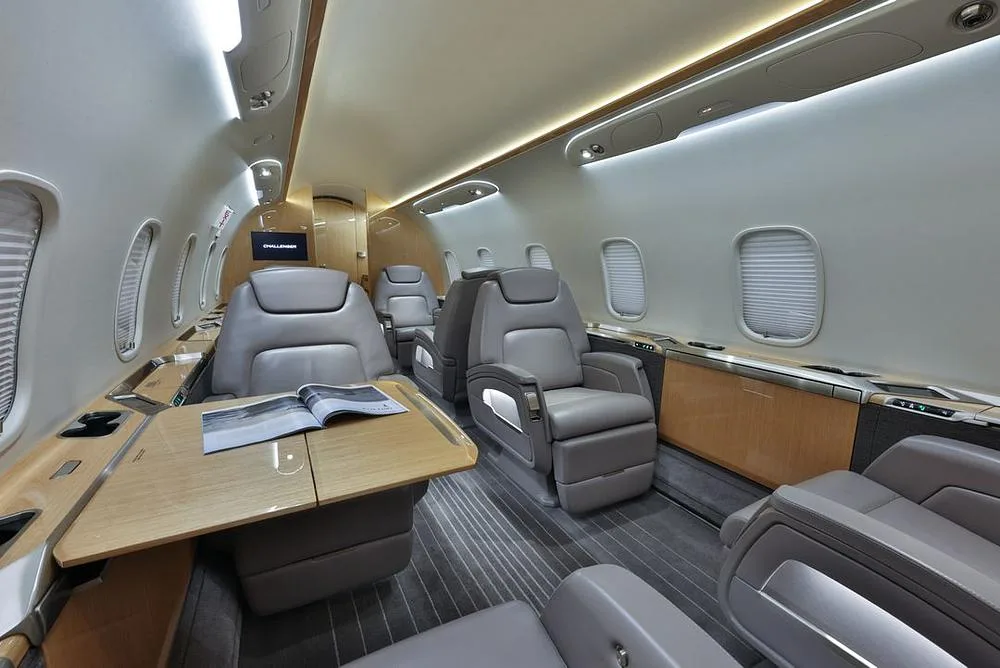 The luxurious interior cabin of a business jet, featuring grey leather seats, a wooden table with a magazine, and overhead lighting.