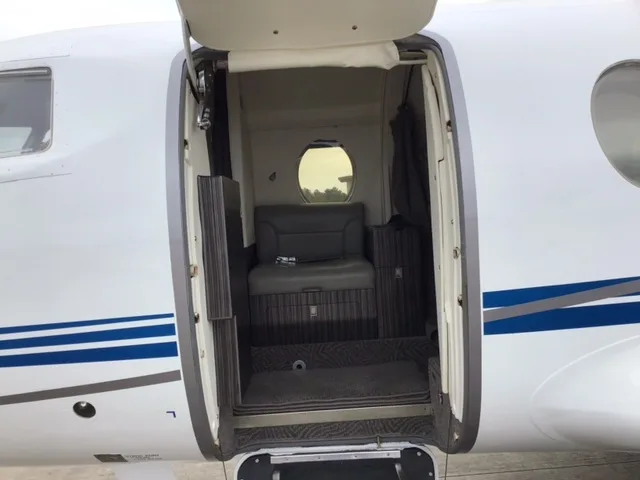 The open entry door of a business jet reveals a small interior area with a single seat and a porthole window.