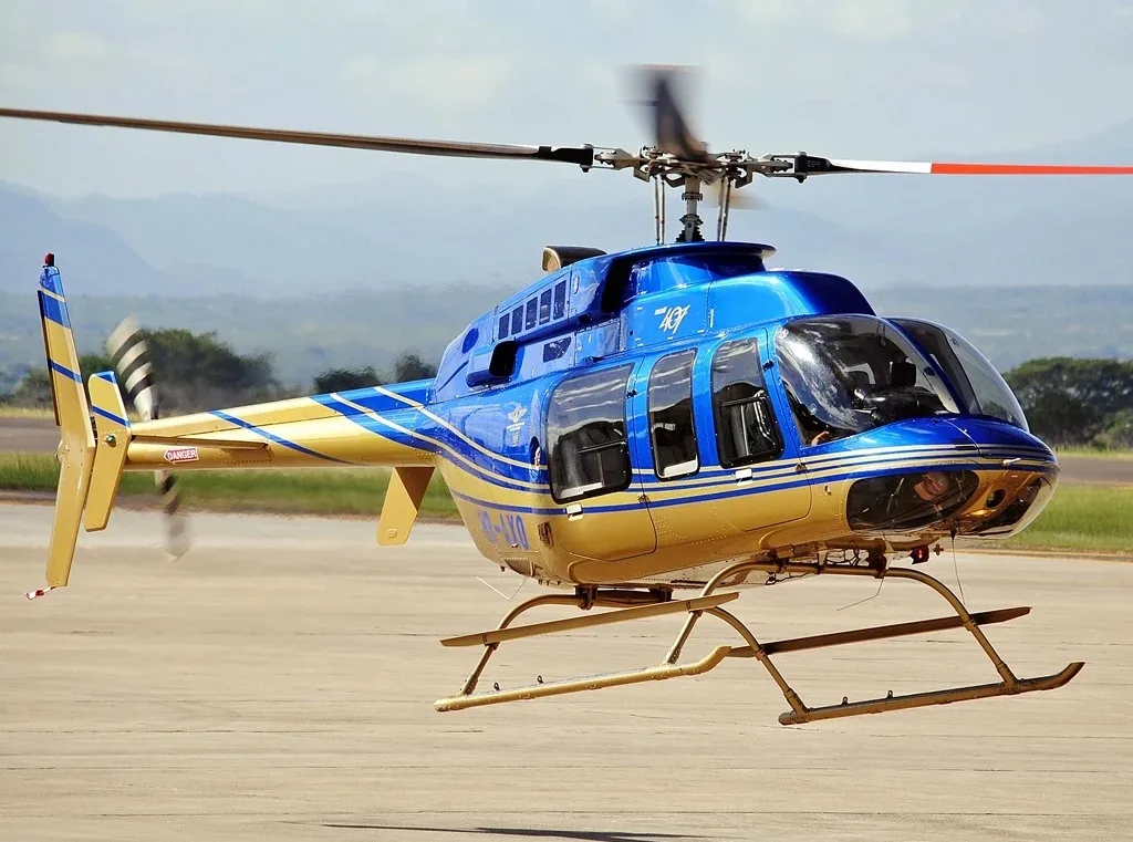 A blue and gold helicopter hovers just above the ground with mountains in the background.