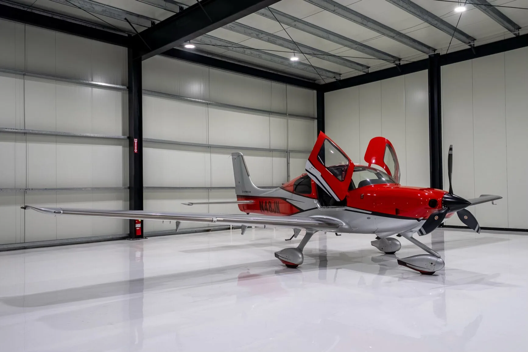 A red and silver Cirrus SR22T Carbon aircraft with registration N48JN and open doors, seen from the front-right in a hangar.
