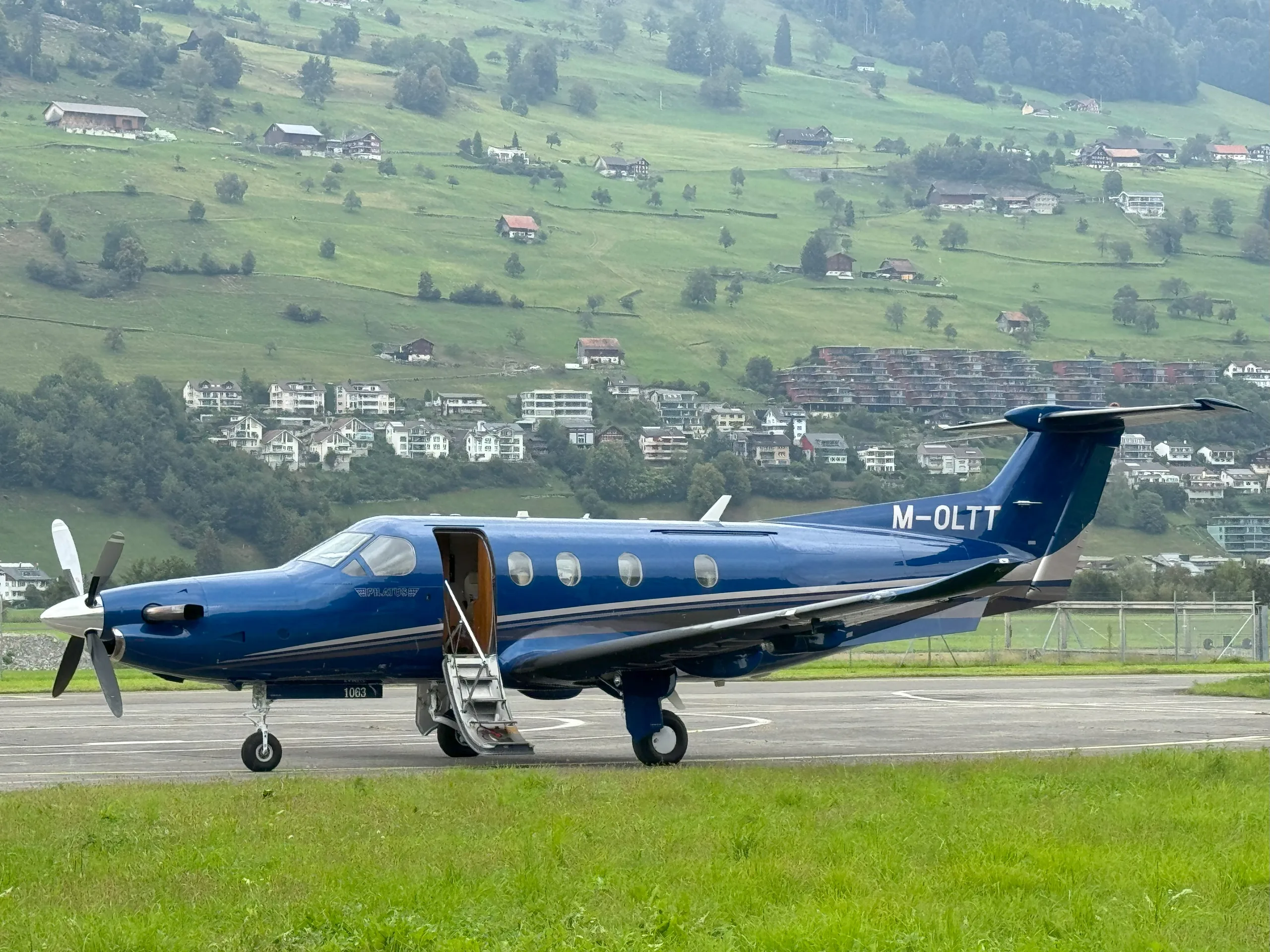 A blue Pilatus PC-12 turboprop aircraft with its door open, parked on an airfield with a scenic mountainous background.