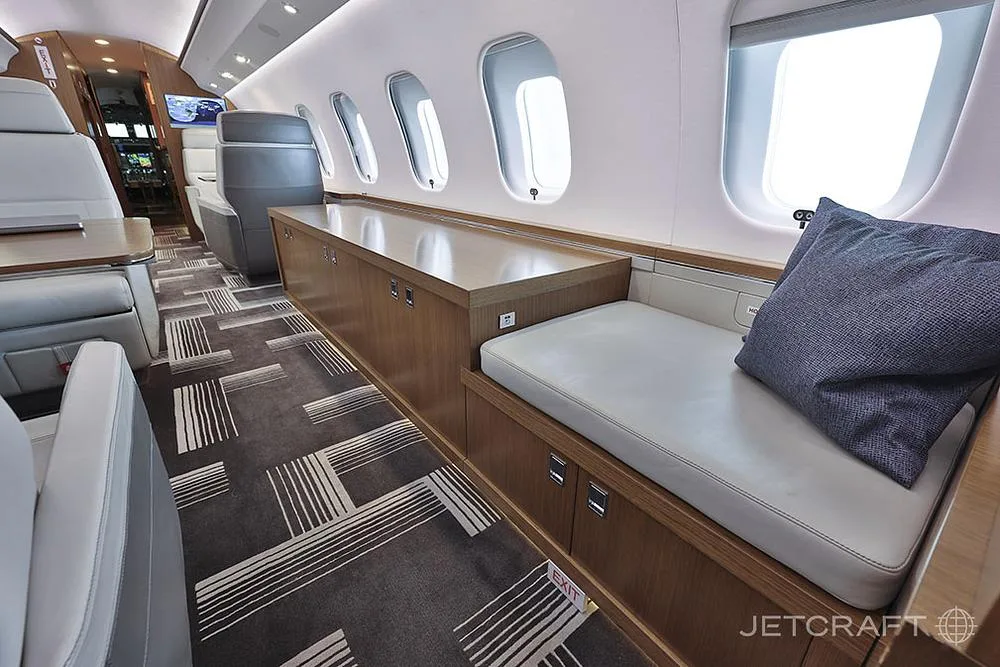 A close-up view of the aircraft cabin showing a wooden credenza, a cushioned bench seat with a pillow, and large windows.