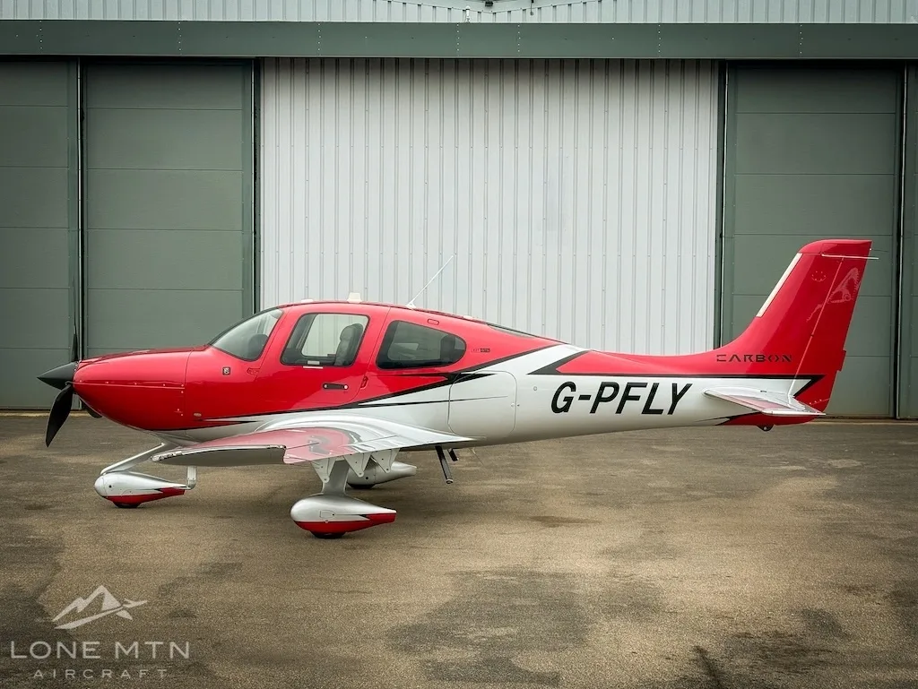 A red and white Cirrus SR22 aircraft (G-PFLY) parked in profile on a tarmac in front of a hangar.