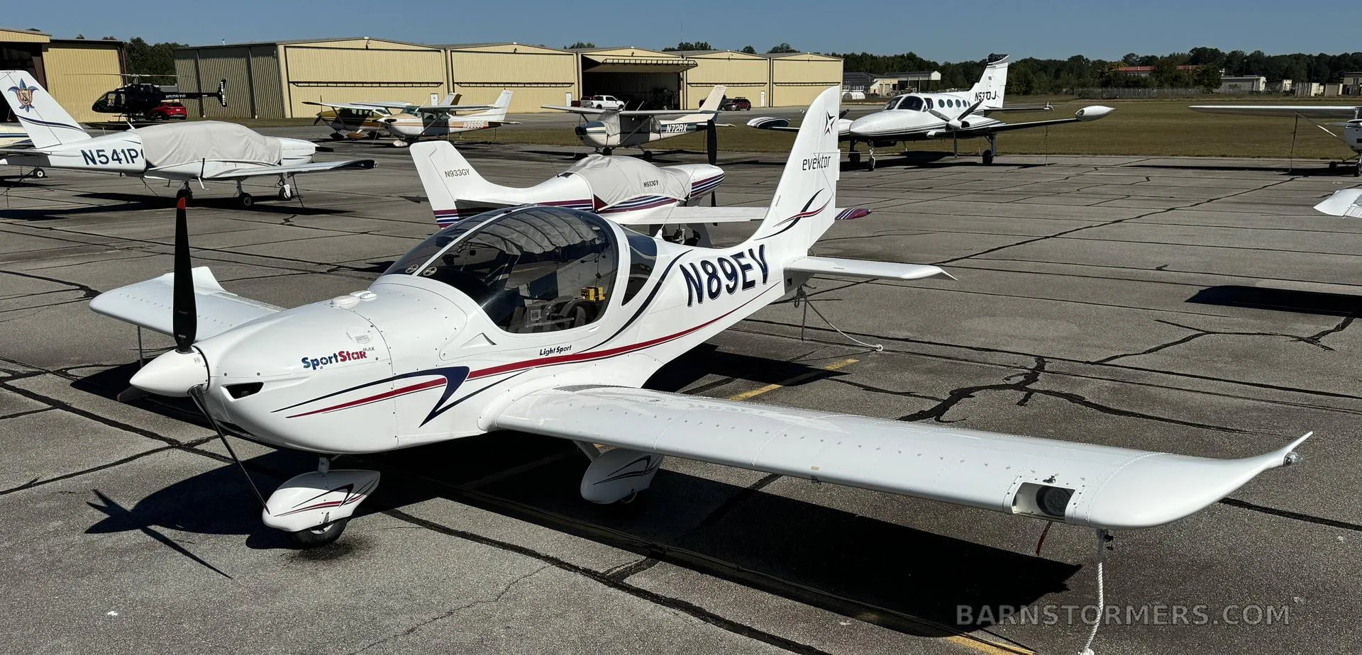 A white Evektor SportStar aircraft (N89EV) with red and blue stripes parked on an airfield, viewed from the front-right.
