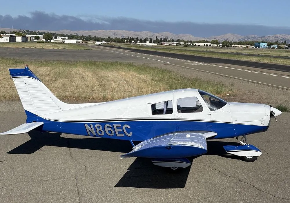 A white and blue Piper Cherokee aircraft (N86EC) parked on an airfield with a runway and distant hills.