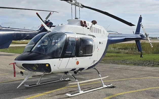 A white and blue helicopter with black accents, parked on an asphalt surface with another helicopter in the background.