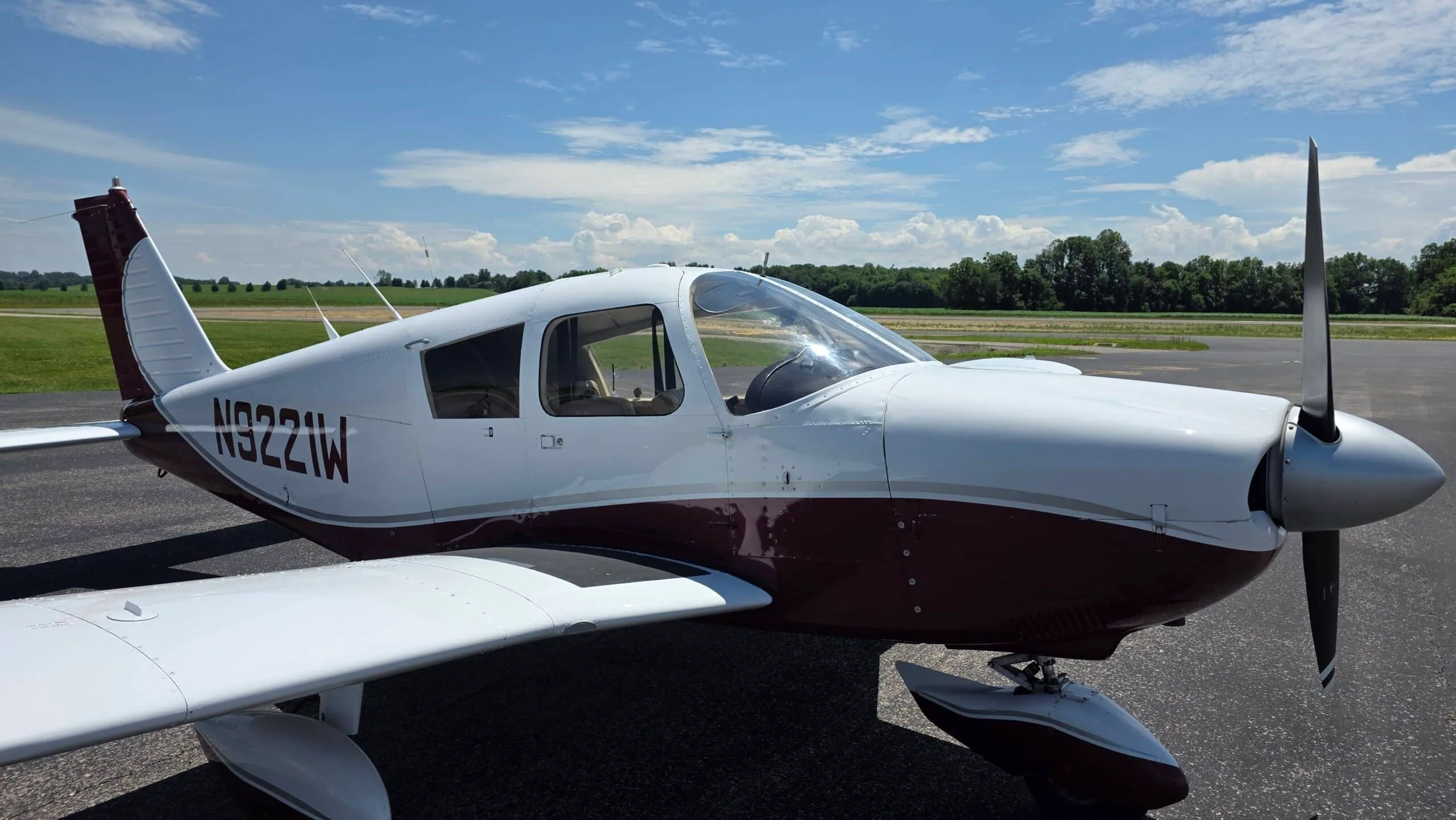 A close-up view of the front right side of a white and maroon Piper Cherokee (N9221W) on an airfield.