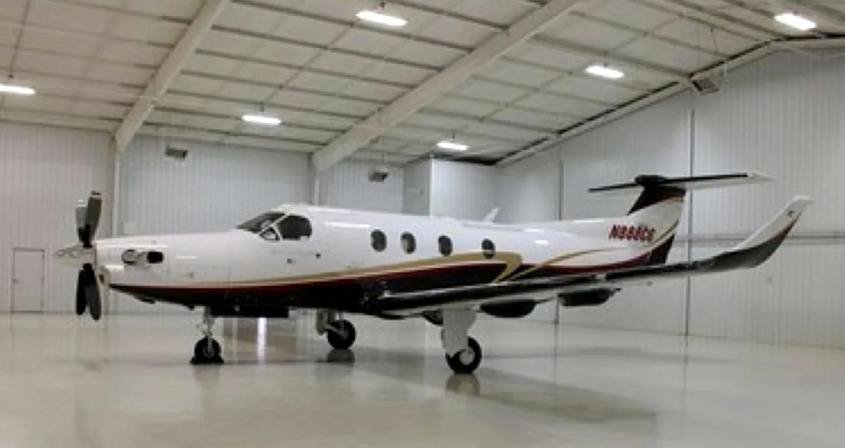 A white Pilatus PC-12 turboprop aircraft with black and gold trim is parked inside a hangar.