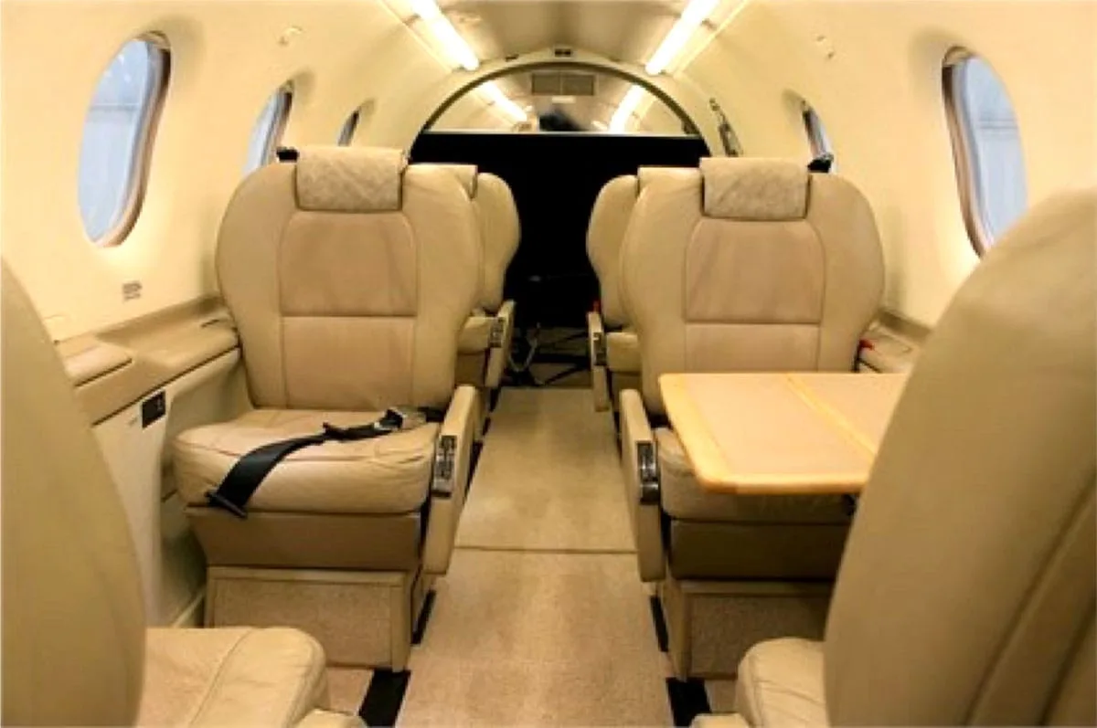 The aircraft cabin interior shows beige leather seats and a small wooden table extended between two seats.