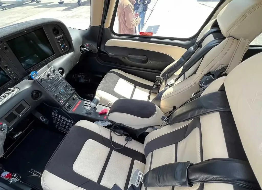 The cockpit and front cabin interior of a small aircraft with beige and black seats and a modern glass cockpit display.