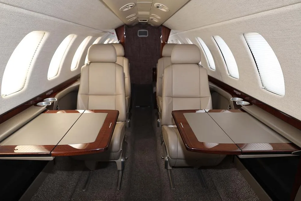The aircraft's interior cabin features beige leather seats, wood trim, and fold-out tables.