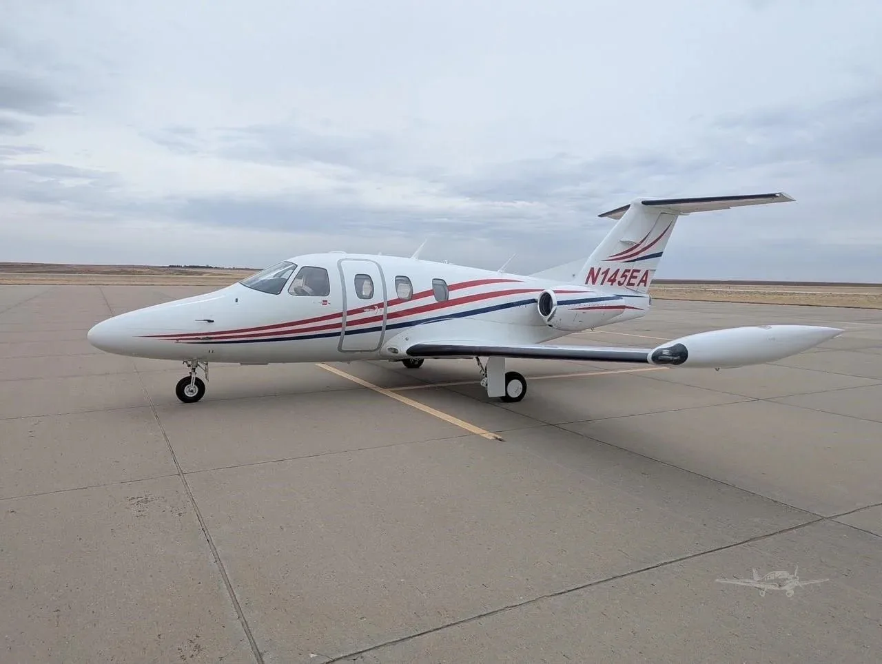 A white Eclipse jet with red and blue stripes, registration N145EA, parked on an airfield under a cloudy sky.