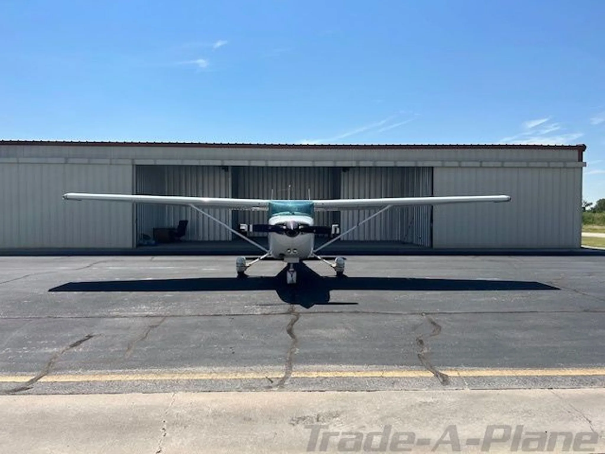 Front view of a white and brown Cessna R172K parked on an asphalt ramp in front of a hangar.