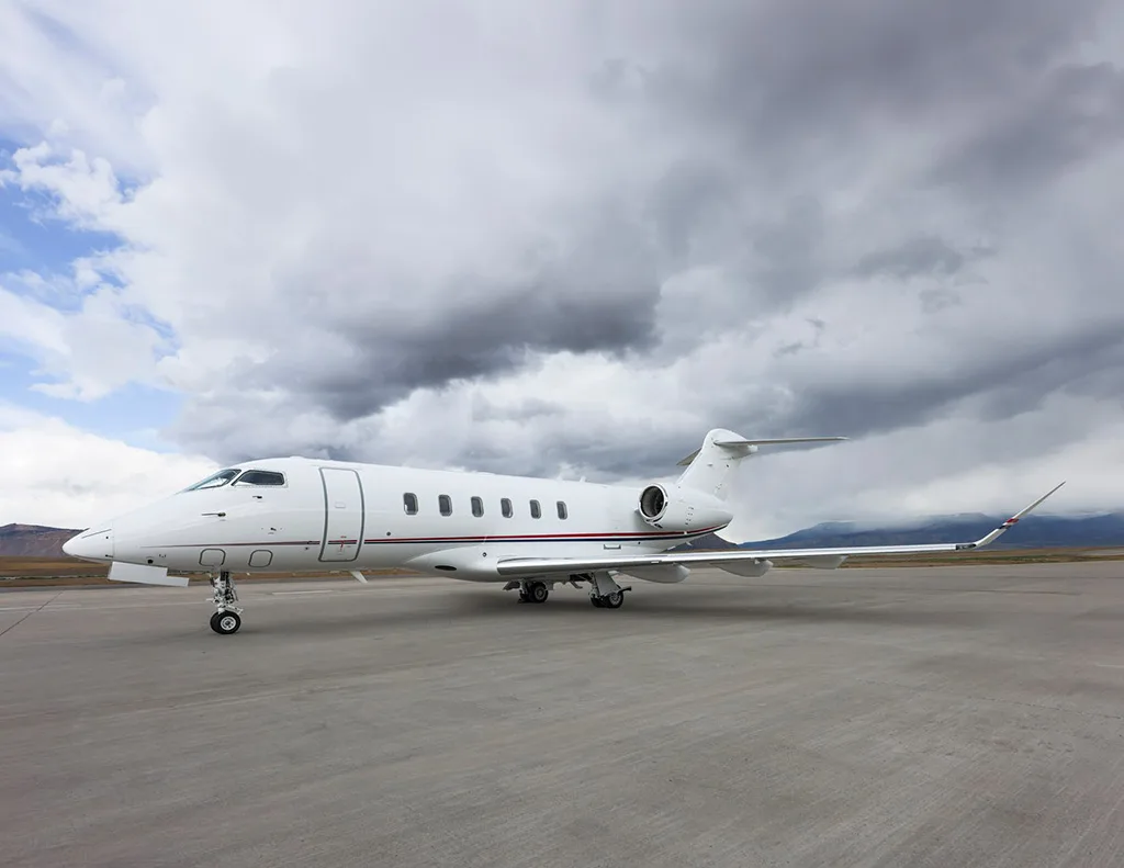 A white Bombardier Challenger 350 business jet with red and blue stripes on the tail, parked on an airfield with mountains in the distance.