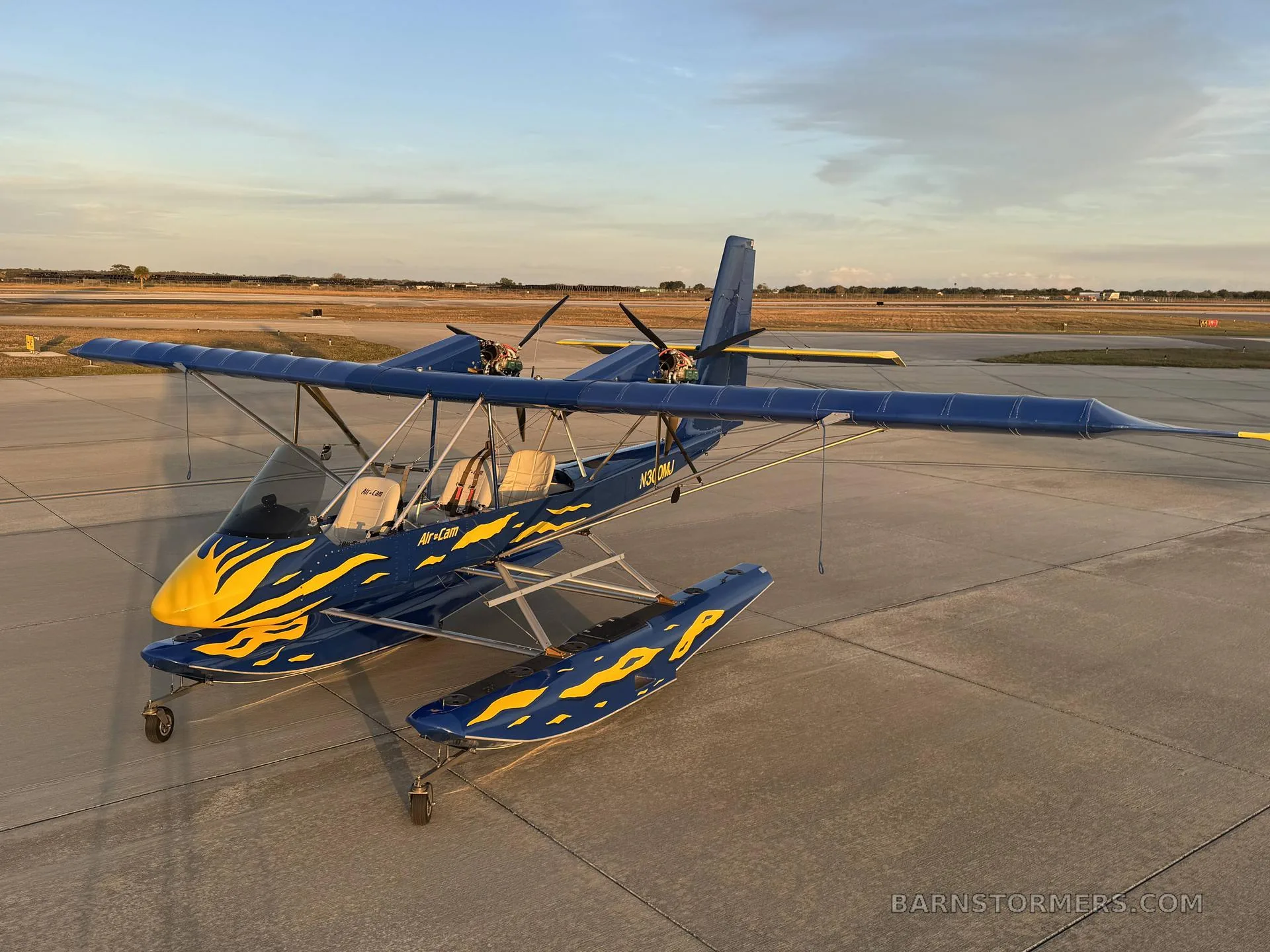 An overhead front-left view of a blue and yellow Air-Cam seaplane with flame decals on an airfield at sunset.