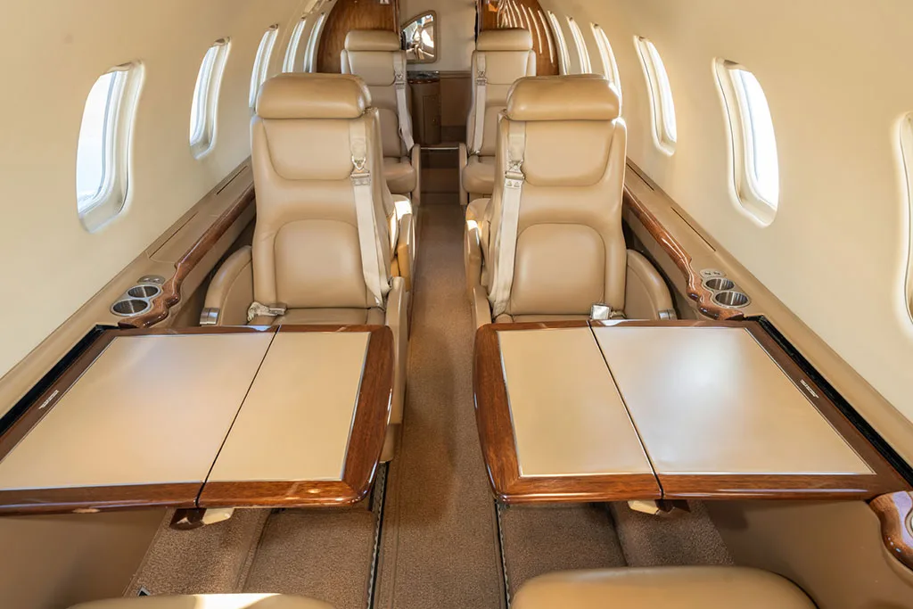 View down the aircraft cabin aisle with beige leather seats and two fold-out tables extended.