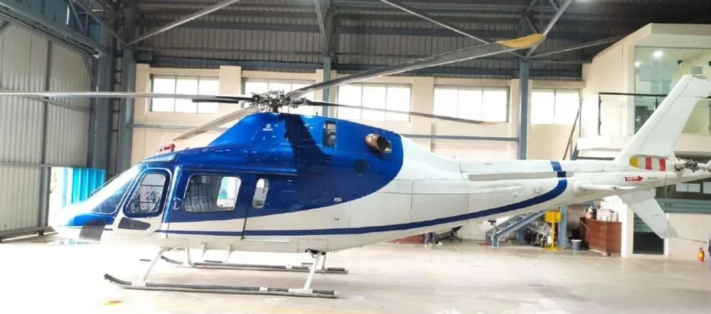 A side view of a blue and white helicopter with a white tail boom, parked inside a hangar.