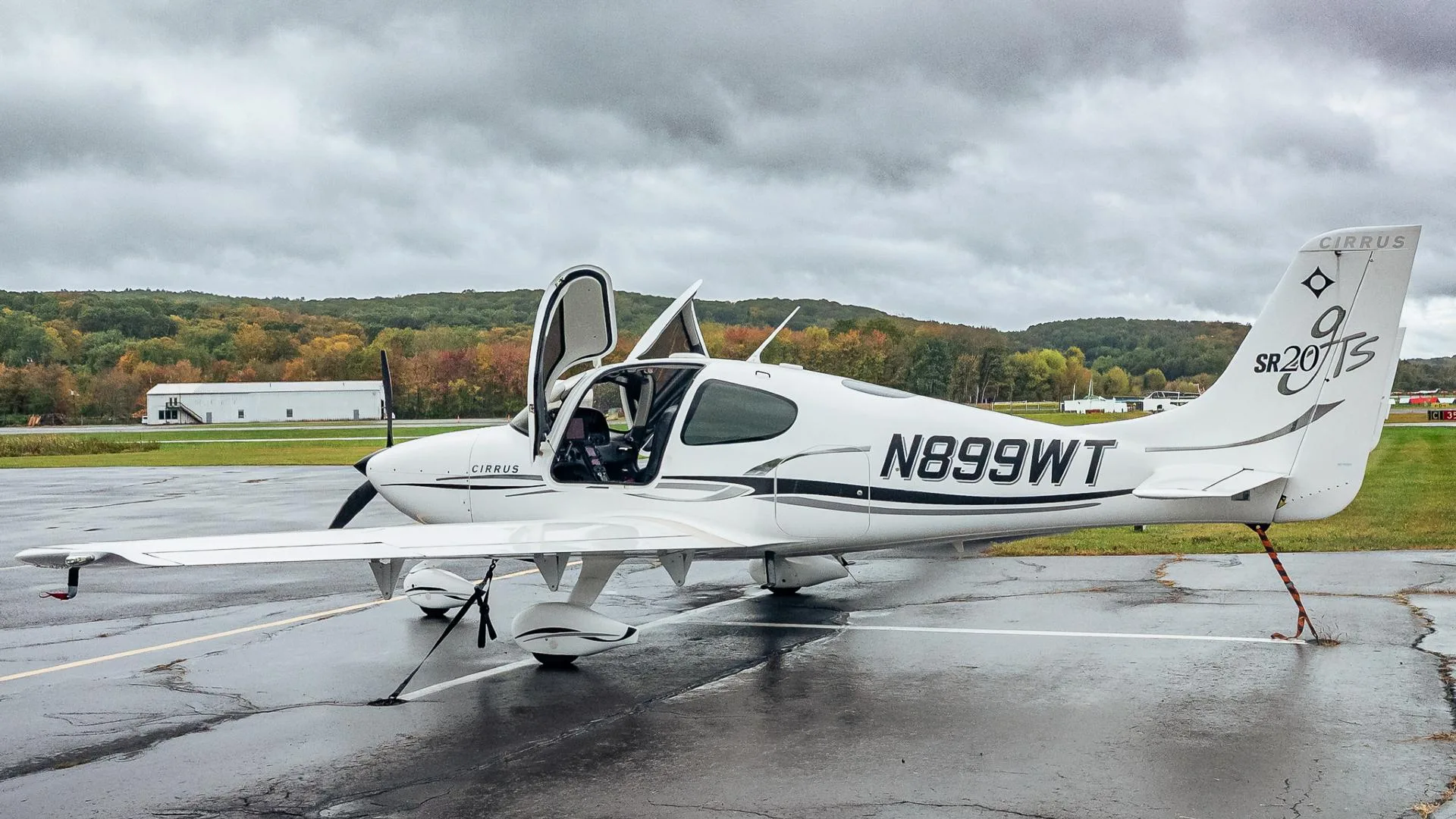 A white Cirrus SR20 aircraft with registration N899WT, parked on a wet tarmac with both gull-wing doors open.