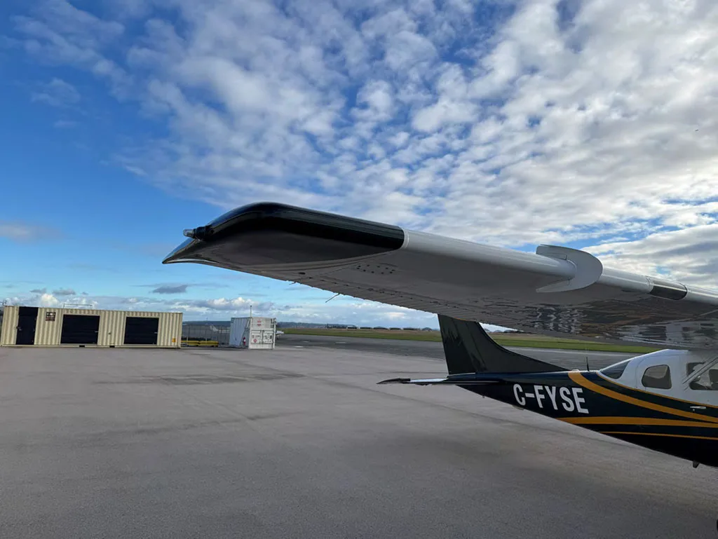 A view from behind the wing of the dark green and white Cessna 182, showing the registration C-FYSE and containers in the background.