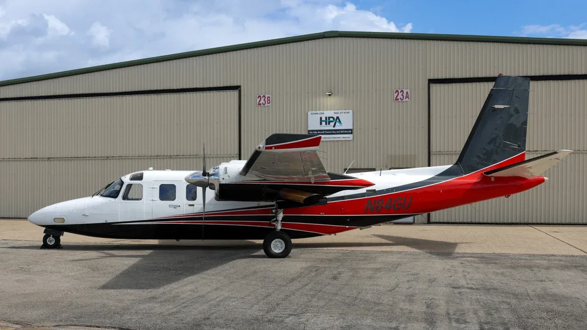 A white, black, and red twin-engine aircraft, N84GU, parked on an tarmac in front of a hangar.