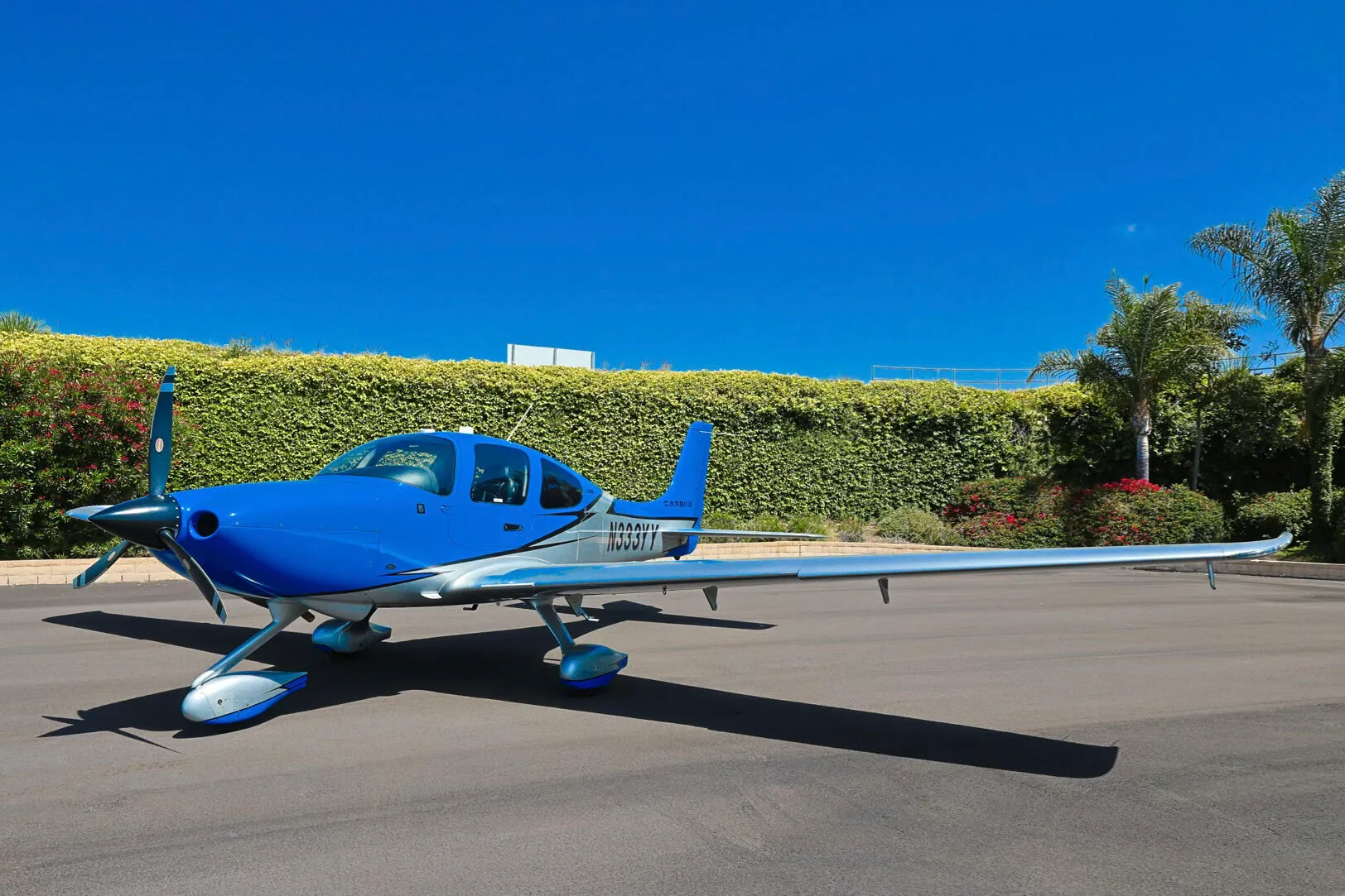 A blue and silver Cirrus SR20 aircraft with tail number N333YY parked on an asphalt ramp with a green hedge in the background.