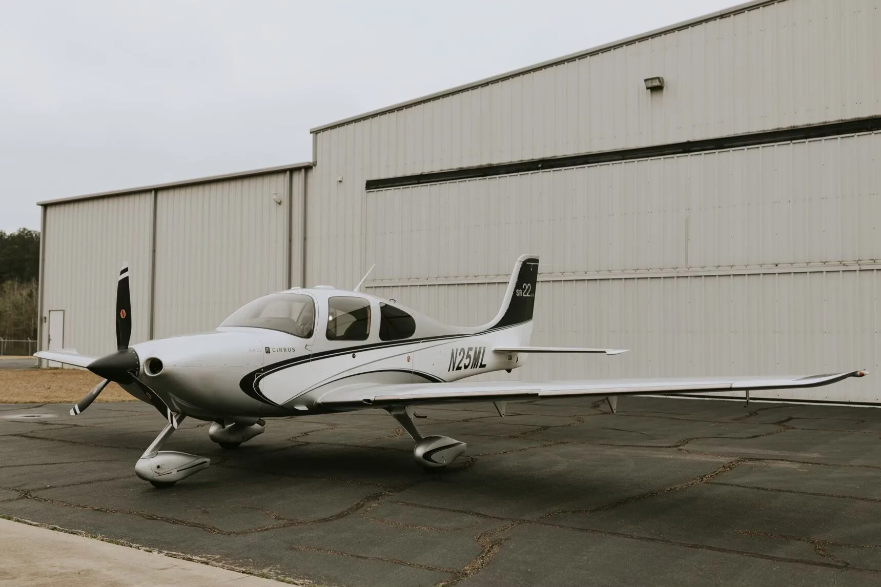 A silver Cirrus SR22 GTS aircraft with black and white stripes parked on an asphalt ramp next to a hangar.