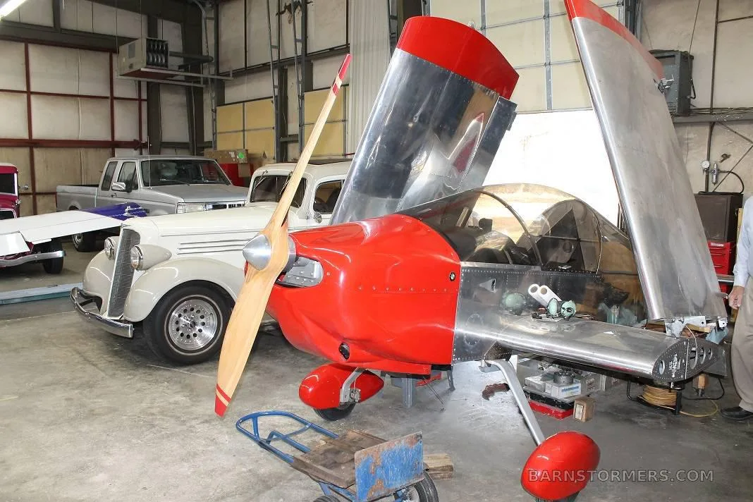 A silver and red experimental aircraft with open wings is parked next to classic cars in a workshop.