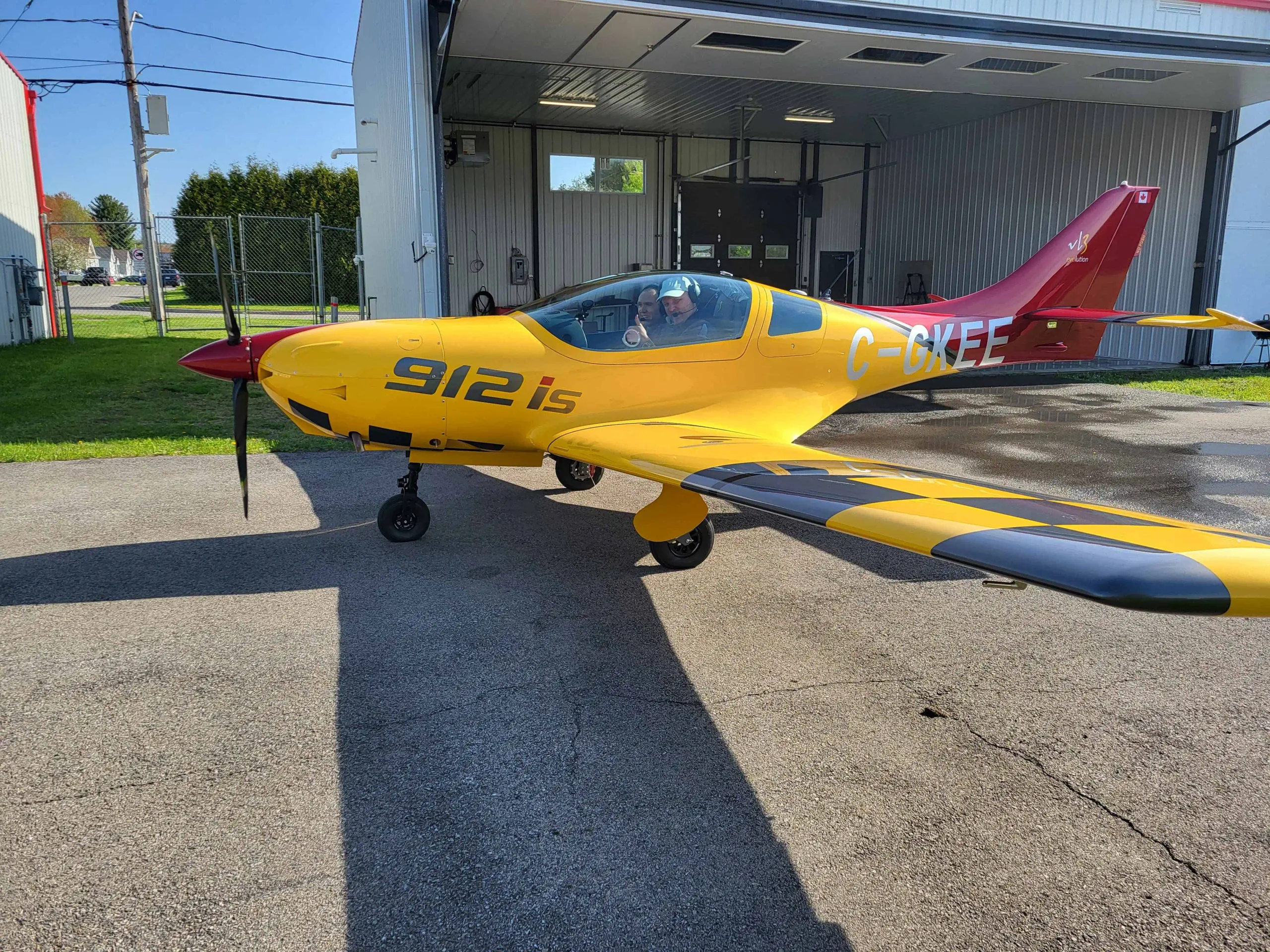 A yellow and red light aircraft, registration C-GKEE, with a checkered wing pattern, parked on an asphalt tarmac in front of an open hangar with two people in the cockpit.