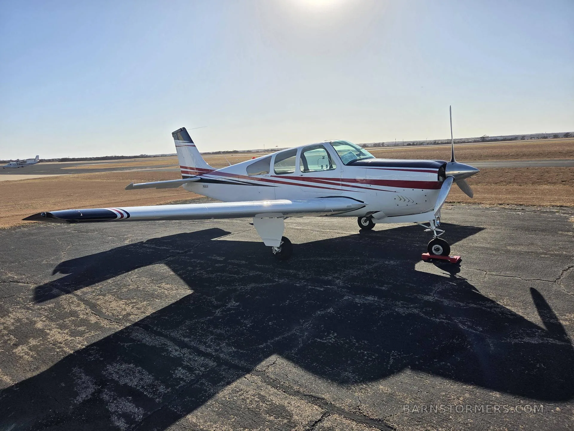 A white and red single-engine aircraft with dark wingtips is parked on an asphalt tarmac under a bright sun.