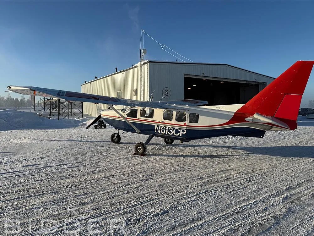 A side view of the N613CP Cessna 206, blue and white with red accents, parked on a snowy airfield next to a white hangar.