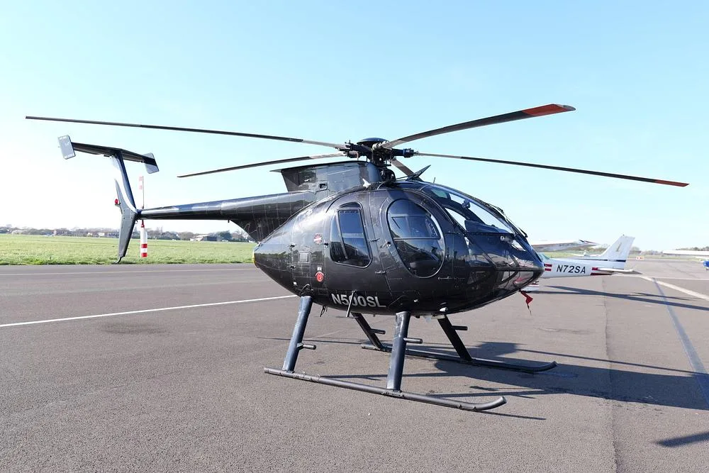 A black MD 500 helicopter (N500SL) is parked on an airfield tarmac, with a white plane (N72SA) in the background.