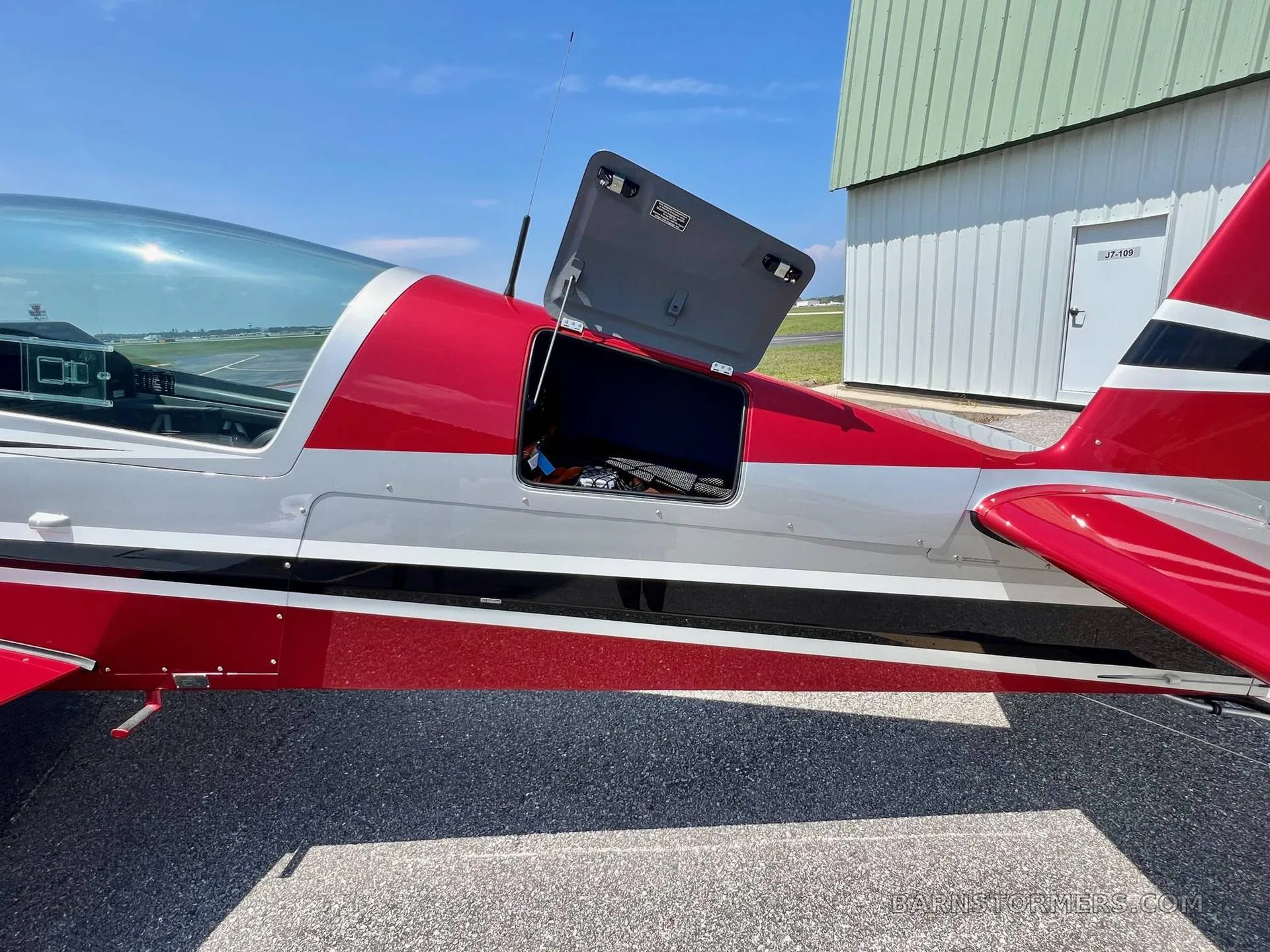 The side of a red and silver aerobatic aircraft shows an open baggage compartment door on the fuselage.