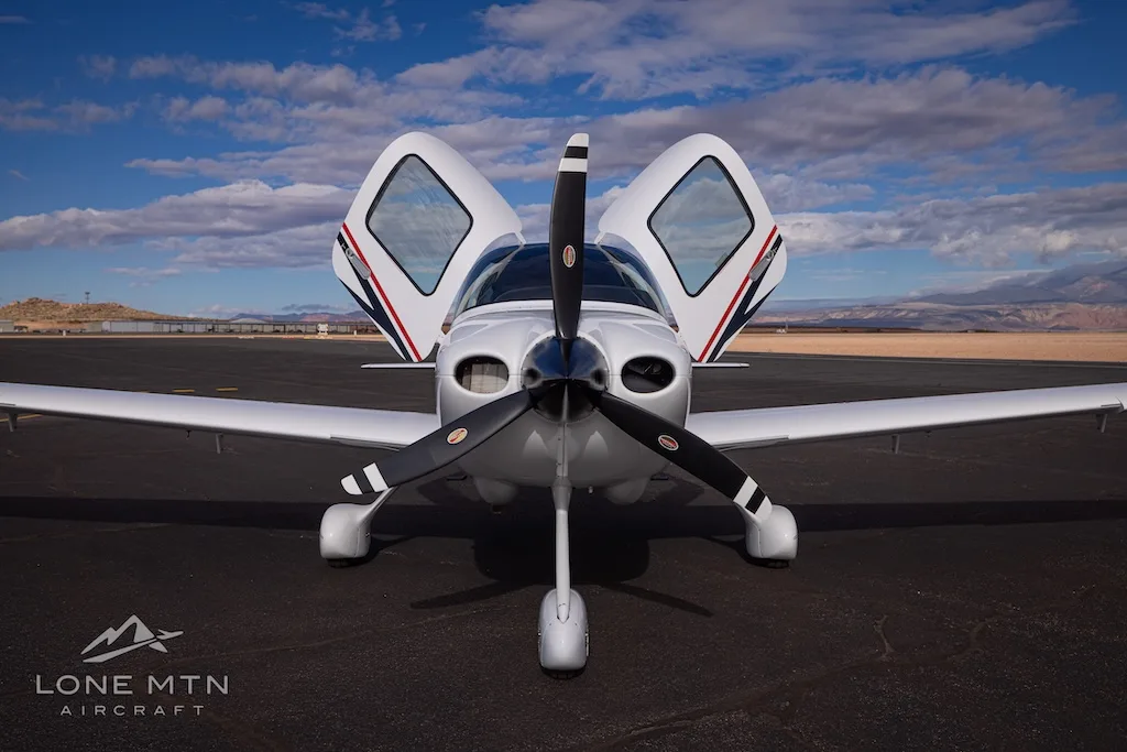 A head-on view of a white Cirrus SR22 aircraft with black and red stripes, registration N418DE, parked on an airfield with doors open.