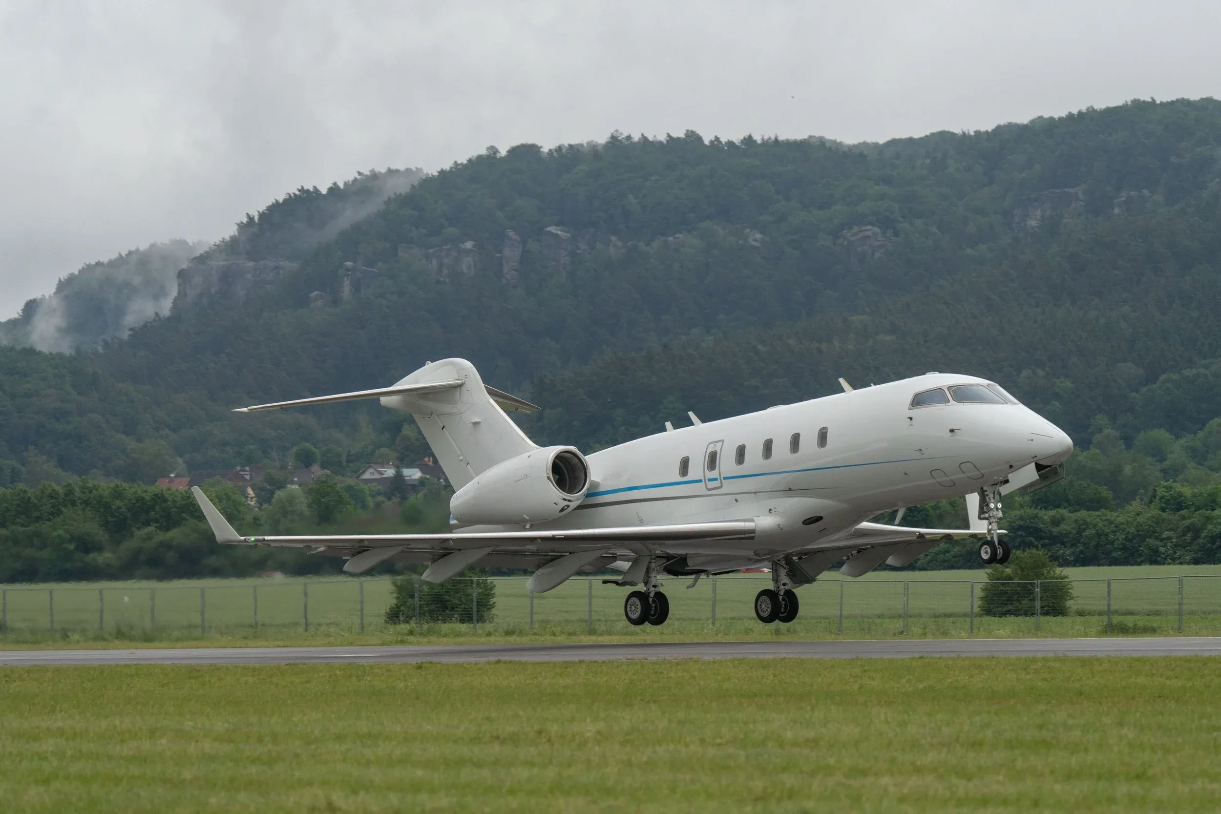 A white business jet with a blue stripe is seen taking off or landing on a runway with green grass and forested hills in the background.