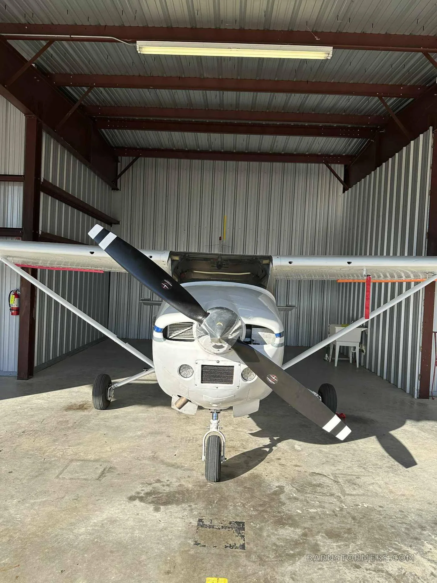 A white single-engine propeller aircraft with blue stripes, seen from the front in a hangar, with its engine cowling open.