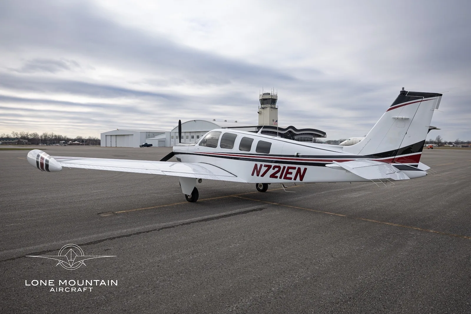 A white and black single-engine propeller aircraft, N721EN, parked on an airfield with a control tower and hangars in the background.