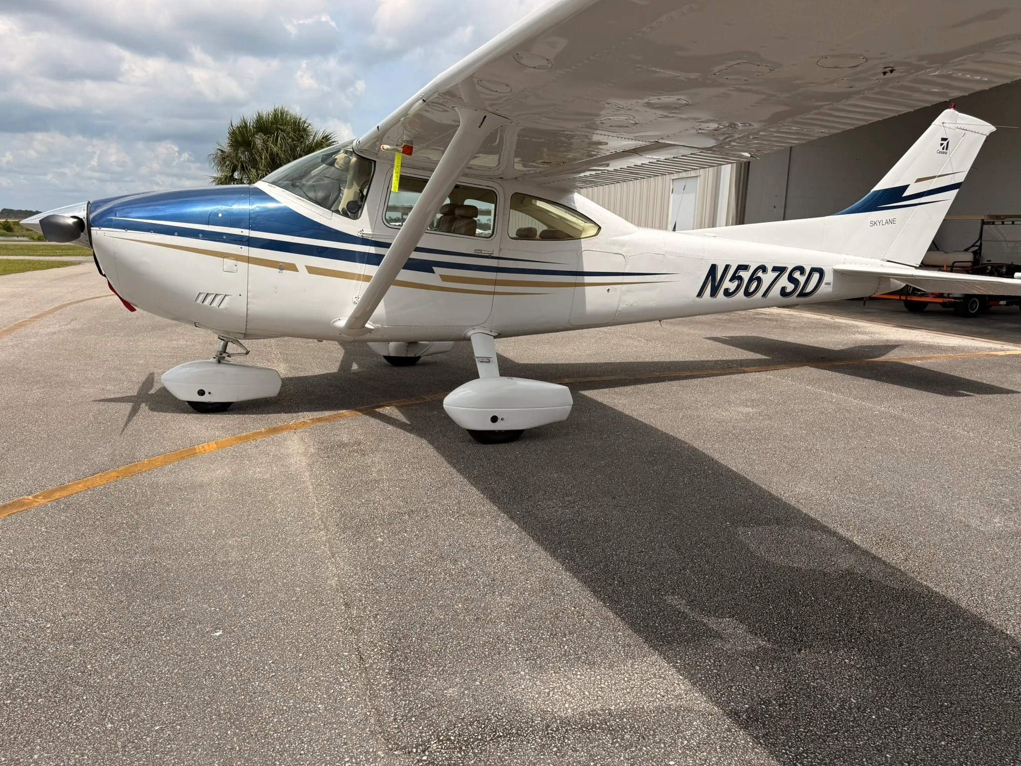A white Cessna Skylane N567SD with blue and gold stripes parked on an asphalt ramp next to a hangar.