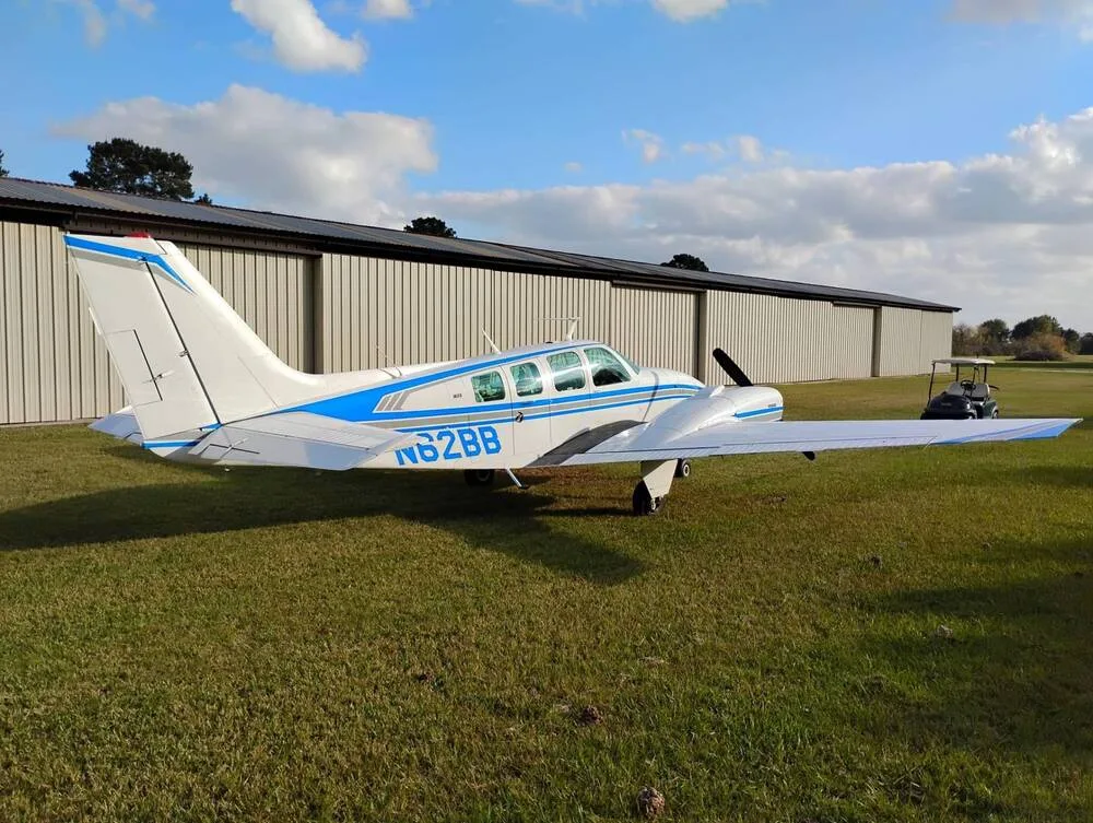 A white Beechcraft Baron N62BB with blue stripes is parked on a grassy field next to a hangar, viewed from the rear-left.