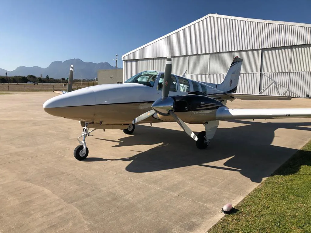 A white and gold twin-engine propeller aircraft parked on a concrete ramp in front of a metal hangar under a clear blue sky.