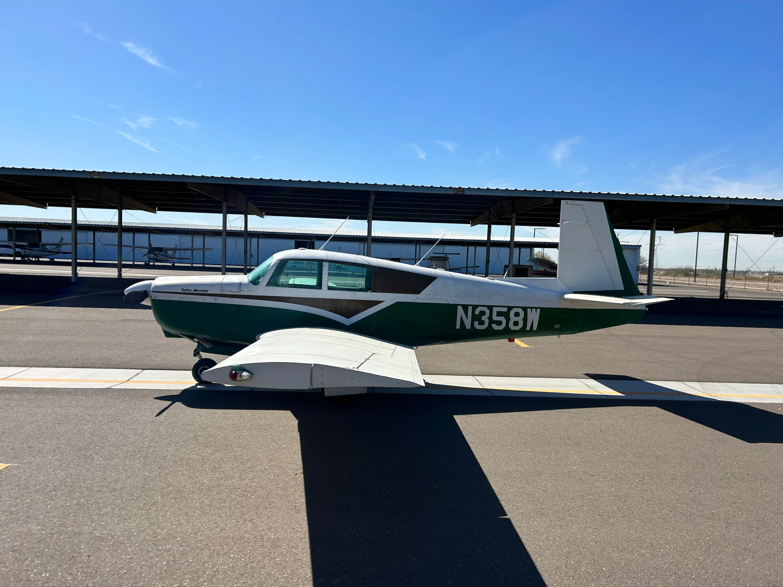 A green and white Turbo Mooney aircraft with registration N358W is parked on an asphalt ramp under a hangar on a sunny day.